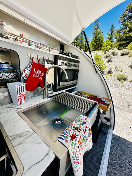 RV rental: compact outdoor kitchen with sink, microwave, "Life is Better at the Campsite" mitt, & nature views.