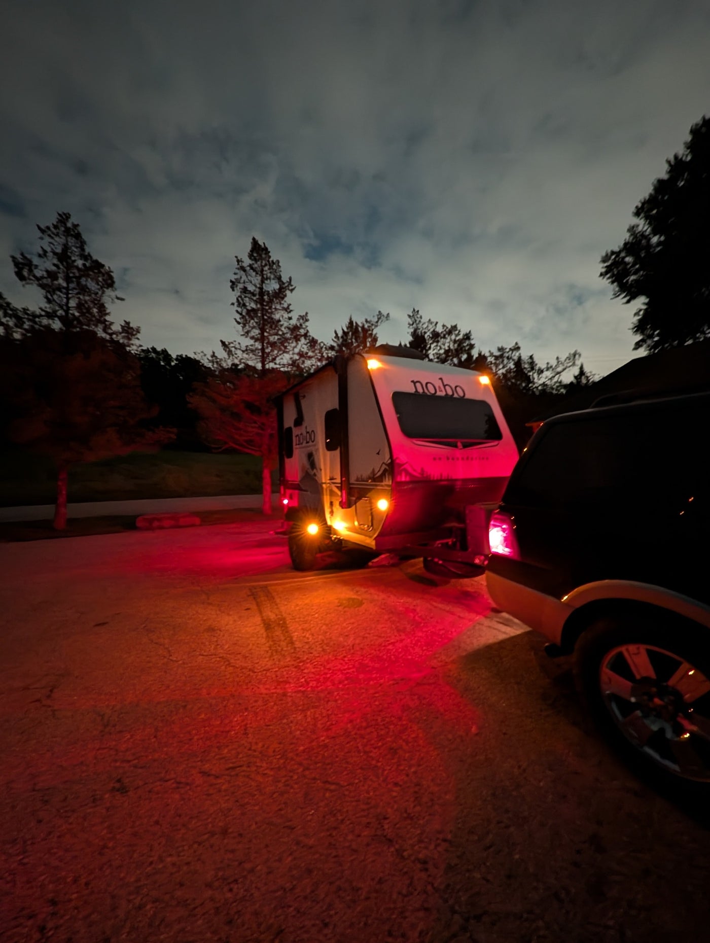 A 'no-bo' travel trailer rv rental, illuminated by its red and amber lights, hitched to a dark SUV at night under a cloudy sky. — photo 20