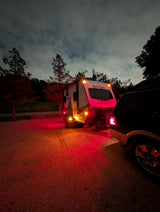 A 'no-bo' travel trailer rv rental, illuminated by its red and amber lights, hitched to a dark SUV at night under a cloudy sky.