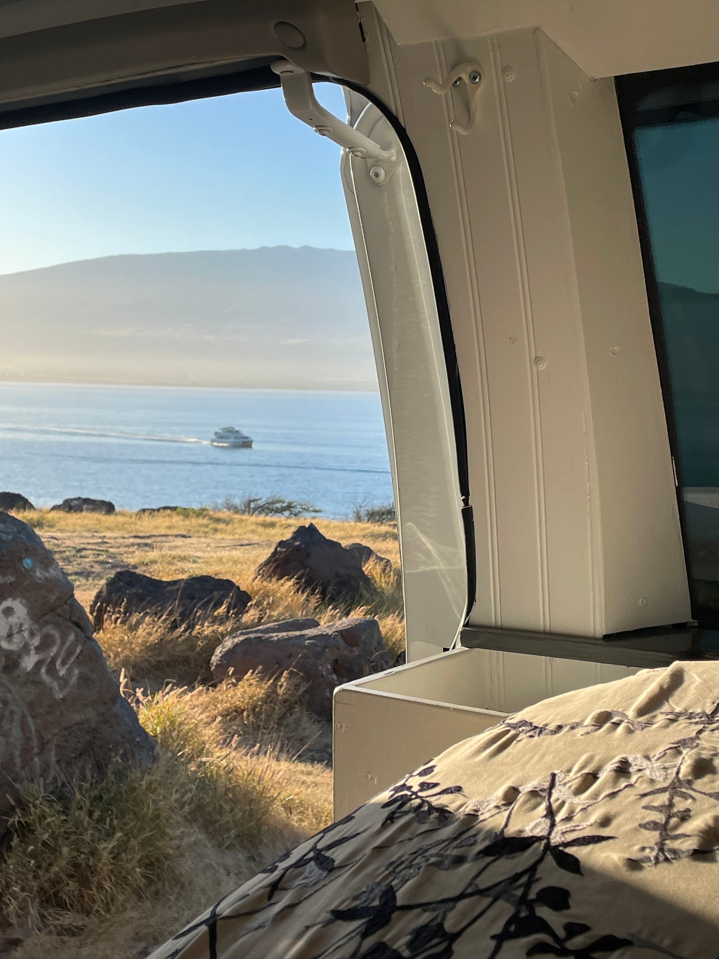 RV rental view: Boat on calm ocean with distant mountains, framed from inside a camper.