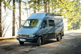 A large, custom gray Mercedes Sprinter camper van for rv rental, parked on a sunlit road in a forest. Perfect for travel adventures.
