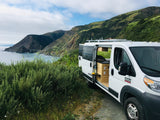 Coastal RV rental dream! White camper van, side door open to reveal a custom interior with pull-out table, overlooking ocean & mountains.