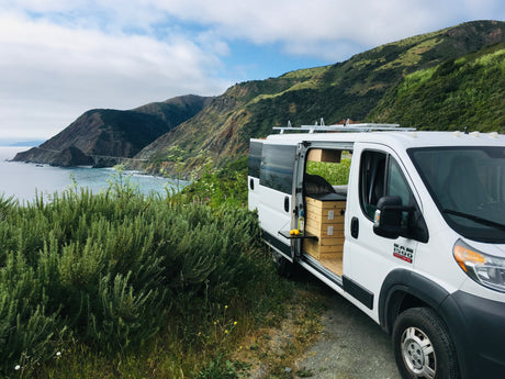 Coastal RV rental dream! White camper van, side door open to reveal a custom interior with pull-out table, overlooking ocean & mountains.