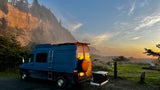 Blue RV rental parked at sunset by a vast, misty coastal cliff. A campfire glows in the grassy field nearby.