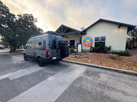 Customized gray RV rental van with roof rack and ladder, parked at Sunset Grill New American Bistro for a travel adventure.