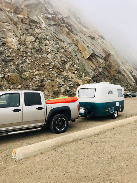 Explorer's RV rental: Truck with kayaks and compact camper trailer parked against a large, misty rocky mountain.