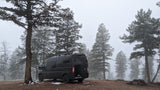 Dark camper van, ideal for rv rental, parked in a serene, snow-dusted pine forest on a foggy winter day.
