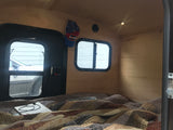 Cozy RV rental camper interior with light wood walls, comfy bed, two windows, and a hanging hat.