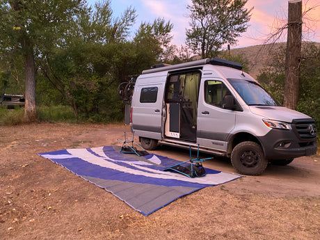 Silver camper van (great for rv rental) parked at a scenic campsite with an outdoor mat and chairs laid out. Enjoy van life!