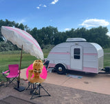 A charming pink & white teardrop camper is set up at a sunny campsite with a matching umbrella. Perfect for your next RV rental adventure!
