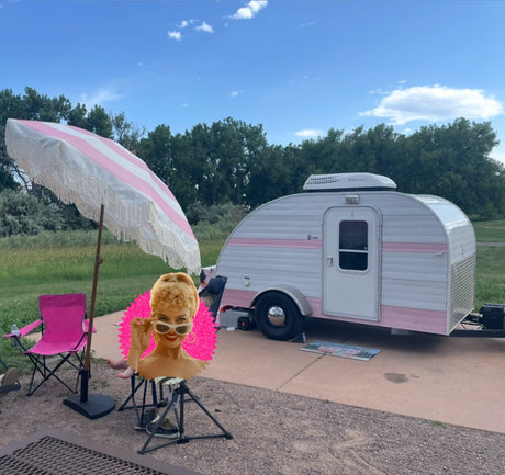 A charming pink & white teardrop camper is set up at a sunny campsite with a matching umbrella. Perfect for your next RV rental adventure!