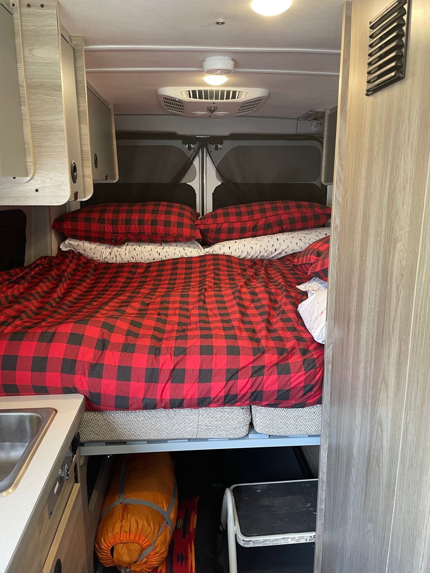 Inside an RV rental: a cozy bed with red plaid bedding, overhead cabinets, and under-bed storage for travel gear.