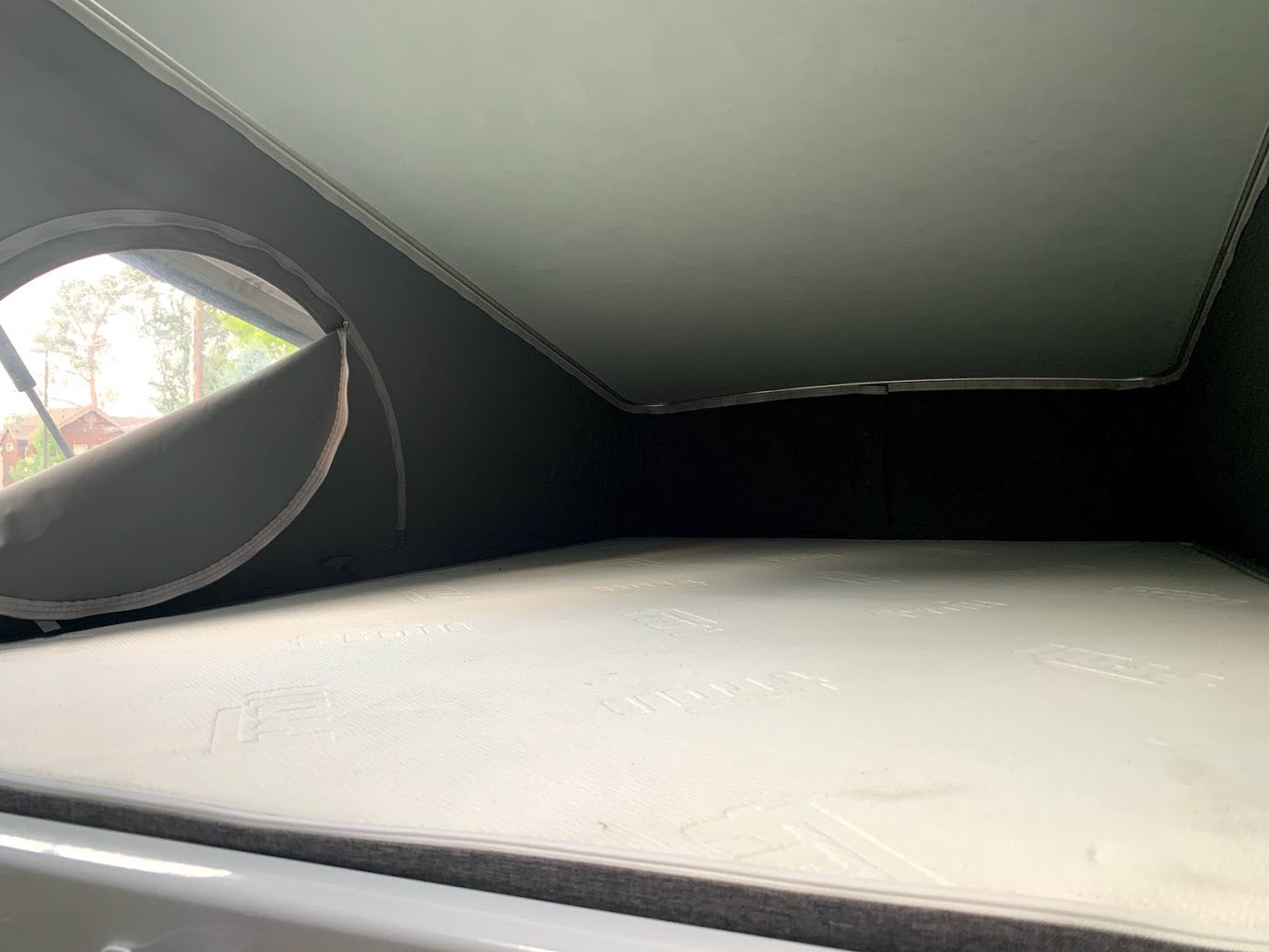 View inside an RV rental's pop-up roof tent, showing a comfortable white mattress and a window with trees outside.