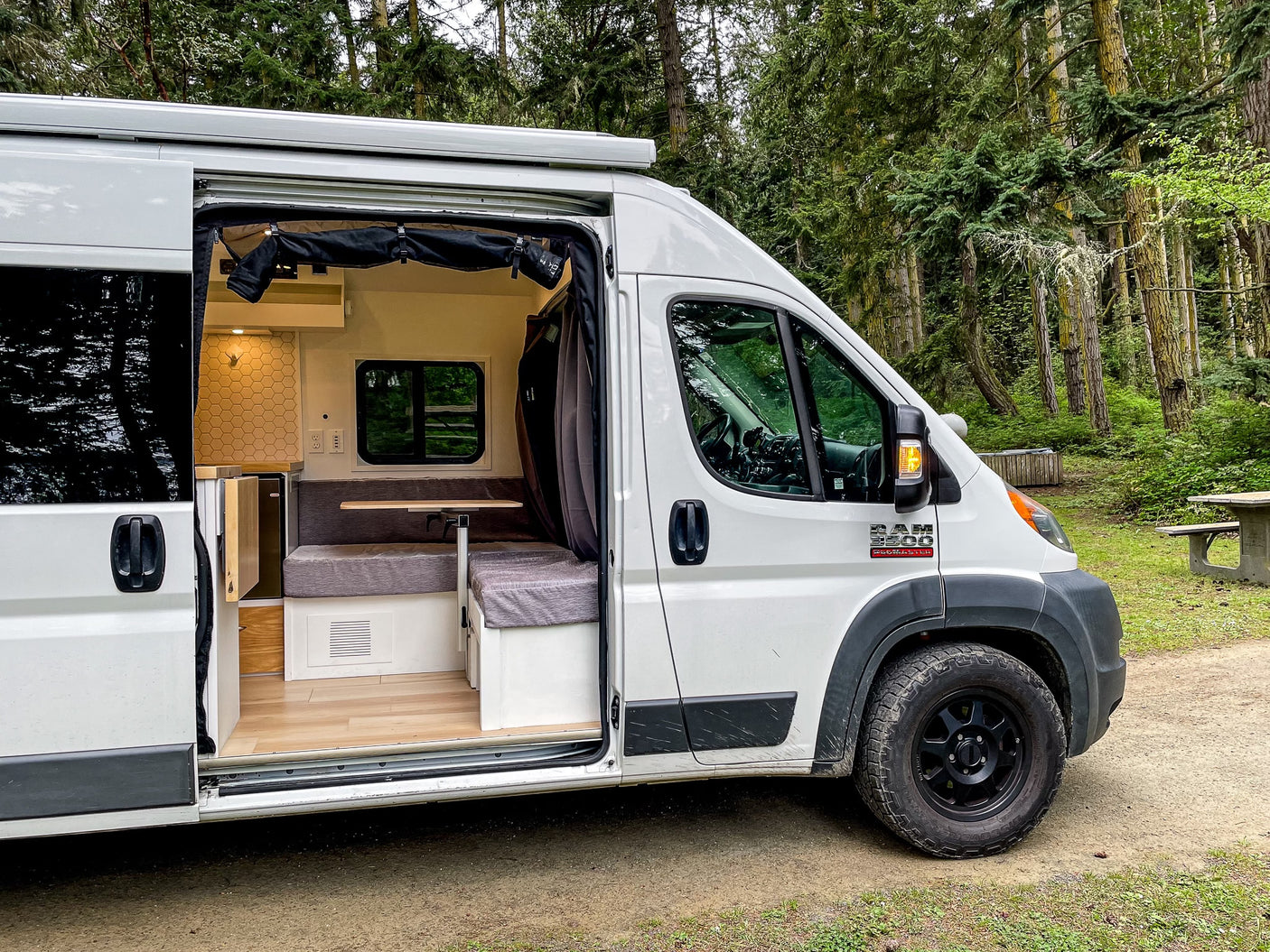 This **rv rental** camper van in a forest reveals its cozy interior, featuring a kitchen, bench seating, and wooden floor.