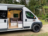 This **rv rental** camper van in a forest reveals its cozy interior, featuring a kitchen, bench seating, and wooden floor.