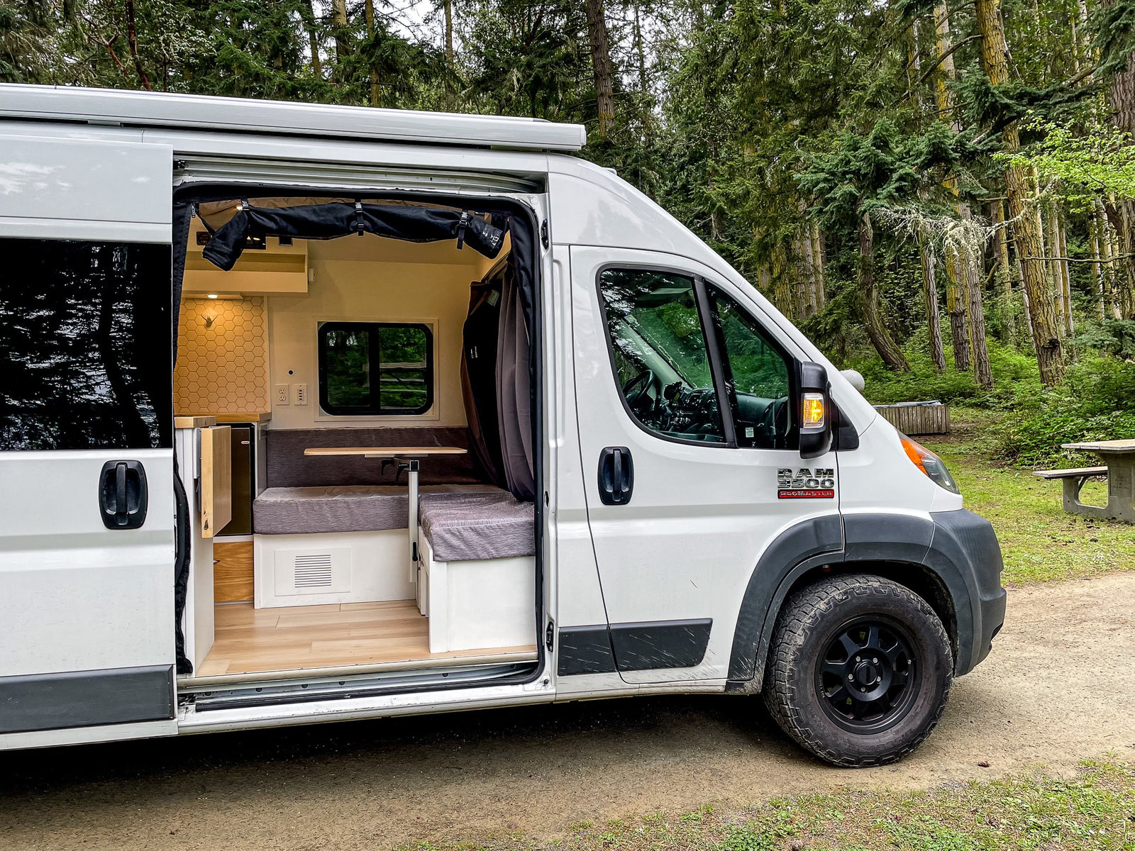 This **rv rental** camper van in a forest reveals its cozy interior, featuring a kitchen, bench seating, and wooden floor.