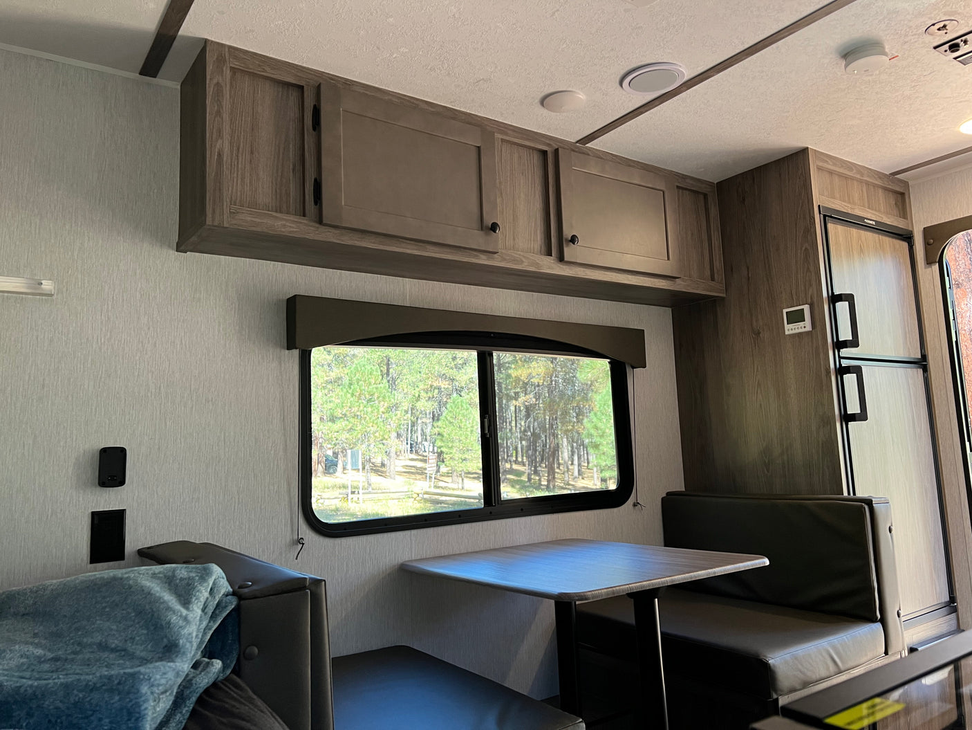 Inside an rv rental: a cozy dinette area with overhead cabinets and a large window overlooking a peaceful wooded campground.