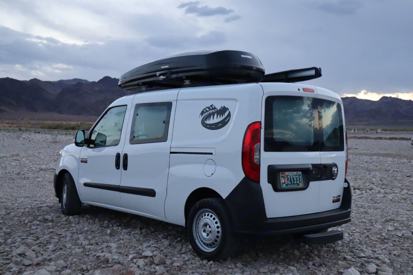 White Ram ProMaster City camper van with roof box, parked on gravel with mountains. Ideal for your next rv rental road trip!