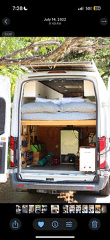 Rear view of an RV rental: a white conversion van with open doors showing an elevated bed, batteries, fridge, and water heater.