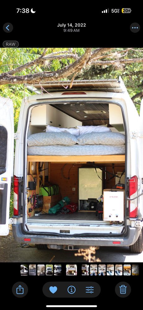Rear view of an RV rental: a white conversion van with open doors showing an elevated bed, batteries, fridge, and water heater.