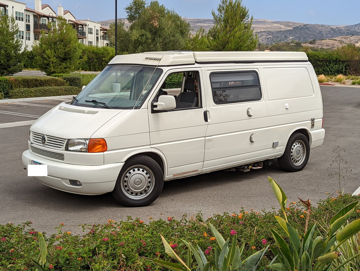 White Winnebago Eurovan camper van, an ideal RV rental, parked on asphalt with buildings and hills in background.