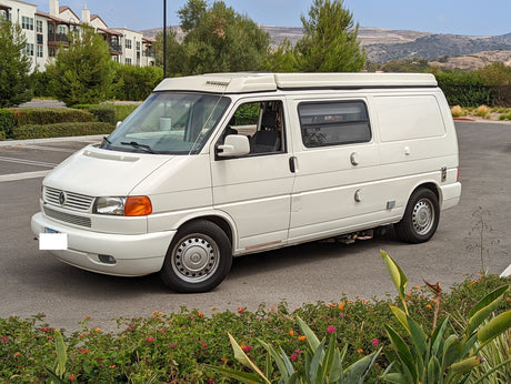 White Winnebago Eurovan camper van, an ideal RV rental, parked on asphalt with buildings and hills in background.