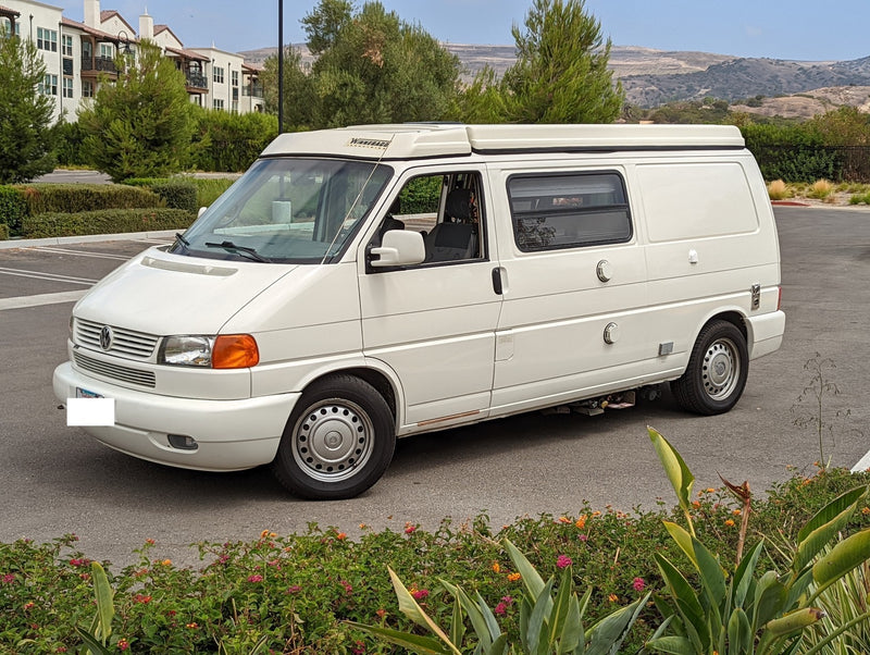 White Winnebago Eurovan camper van, an ideal RV rental, parked on asphalt with buildings and hills in background. — photo 20