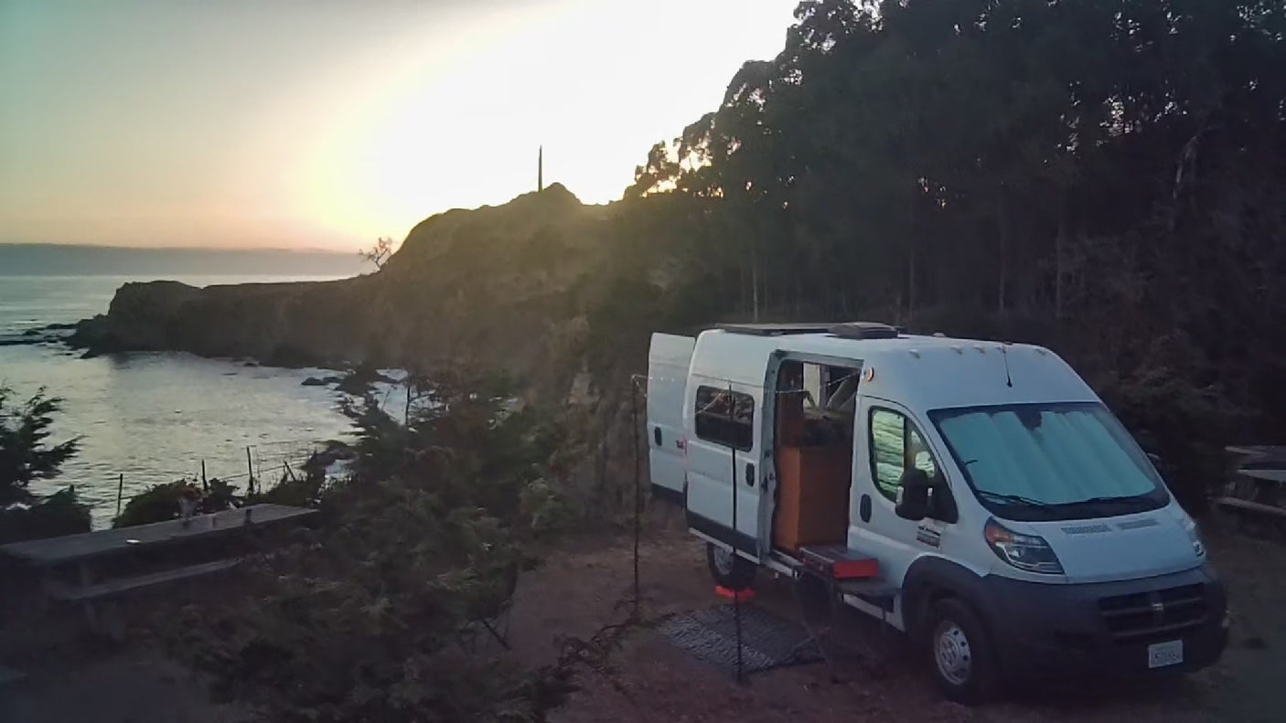 An rv rental van, doors open, parked by cliffs overlooking a sunset ocean view. Perfect coastal camping.