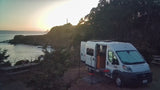 An rv rental van, doors open, parked by cliffs overlooking a sunset ocean view. Perfect coastal camping.