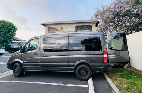 A grey Mercedes Sprinter camper van with a raised pop-top, perfect for an RV rental. Rear doors are open in a sunny lot.