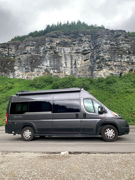 Dark grey camper van RV rental parked on a road beside a massive rocky cliff covered in green foliage and trees.