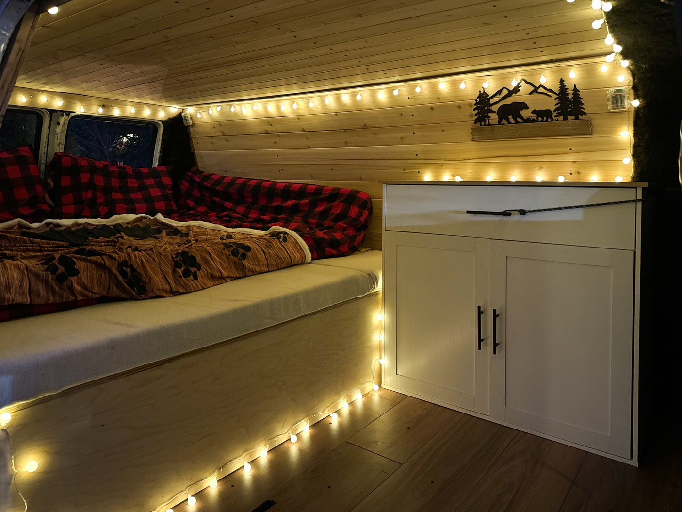 Inside a charming RV rental: rustic wood interior, twinkling lights, comfy plaid bed, and convenient storage unit.