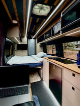 Cozy RV rental interior: laptop workspace, comfy bed, kitchenette with fridge & cooktop. Ideal for travel.