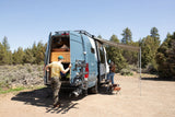 RV rental adventure: A family sets up their van with an awning extended, bikes on back, for camping in nature.