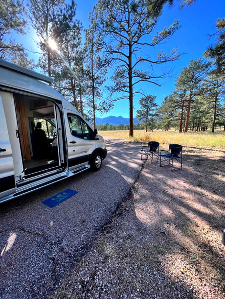 This RV rental offers a sunny outdoor escape. A camper van is parked among pine trees with mountains beyond and chairs set.