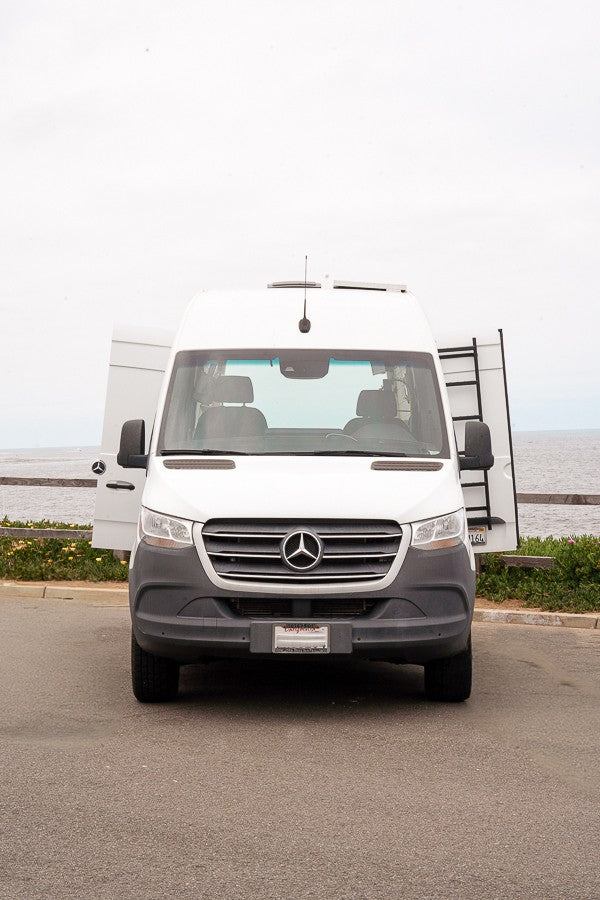 Front view of a white Mercedes-Benz Sprinter rv rental van. Its rear doors are open, revealing a ladder, parked by the ocean.