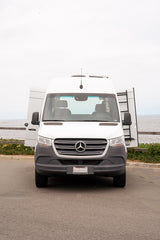 Front view of a white Mercedes-Benz Sprinter rv rental van. Its rear doors are open, revealing a ladder, parked by the ocean.