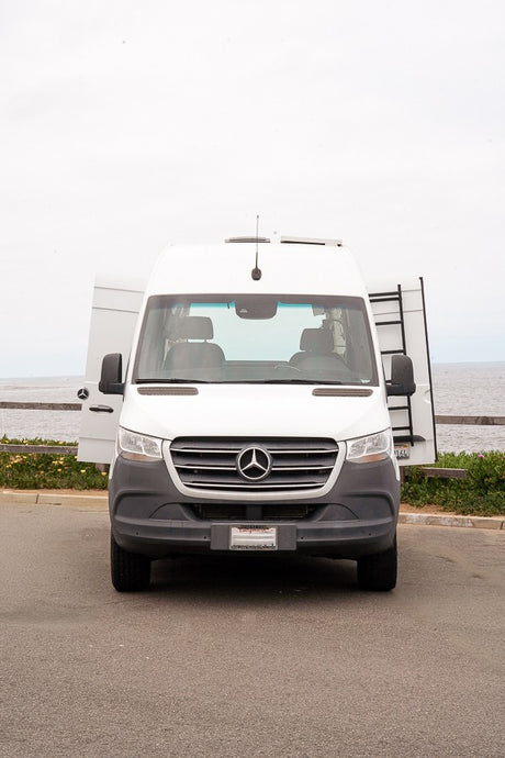 Front view of a white Mercedes-Benz Sprinter rv rental van. Its rear doors are open, revealing a ladder, parked by the ocean.
