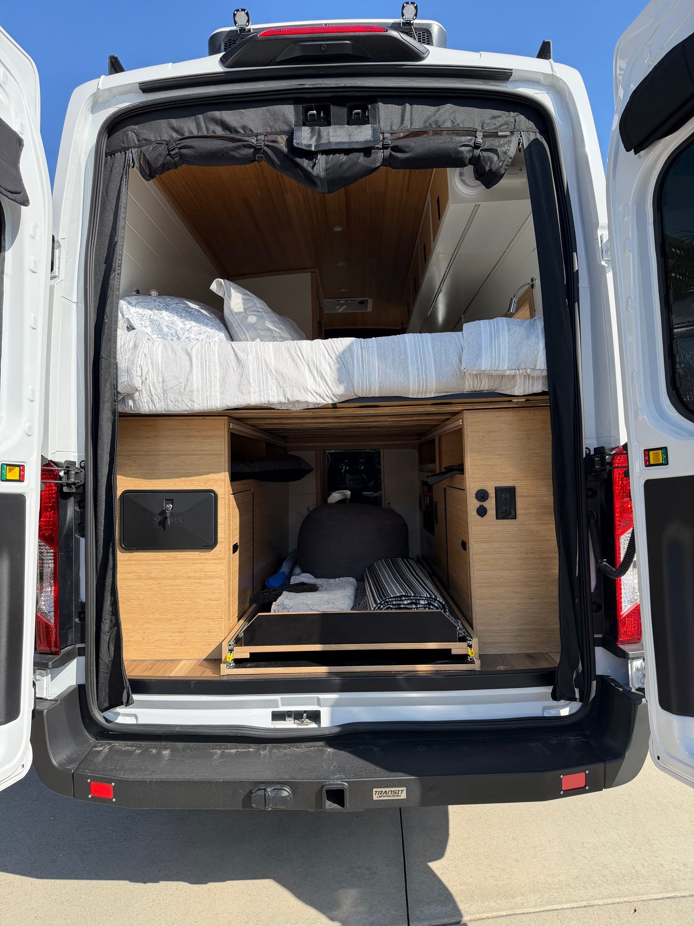 RV rental: Rear view of a white camper van's interior, featuring an elevated bed, custom wood storage, and pull-out tray.