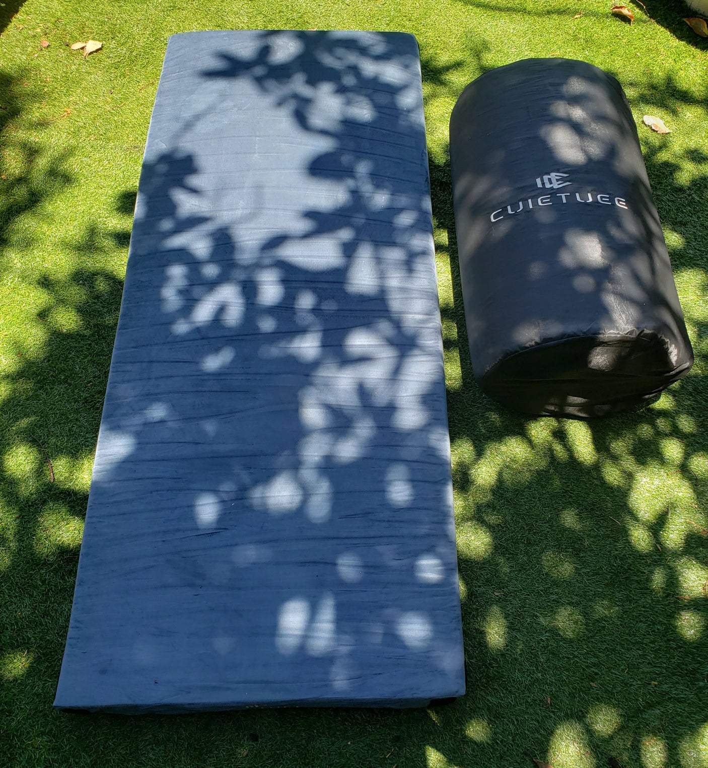 Essential RV rental comfort: a long grey sleeping mat and a black branded cylindrical bag on sun-dappled artificial grass.