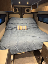 Cozy bed area in an RV rental with blue bedding, overhead storage, speakers, and under-bed appliances and safe.