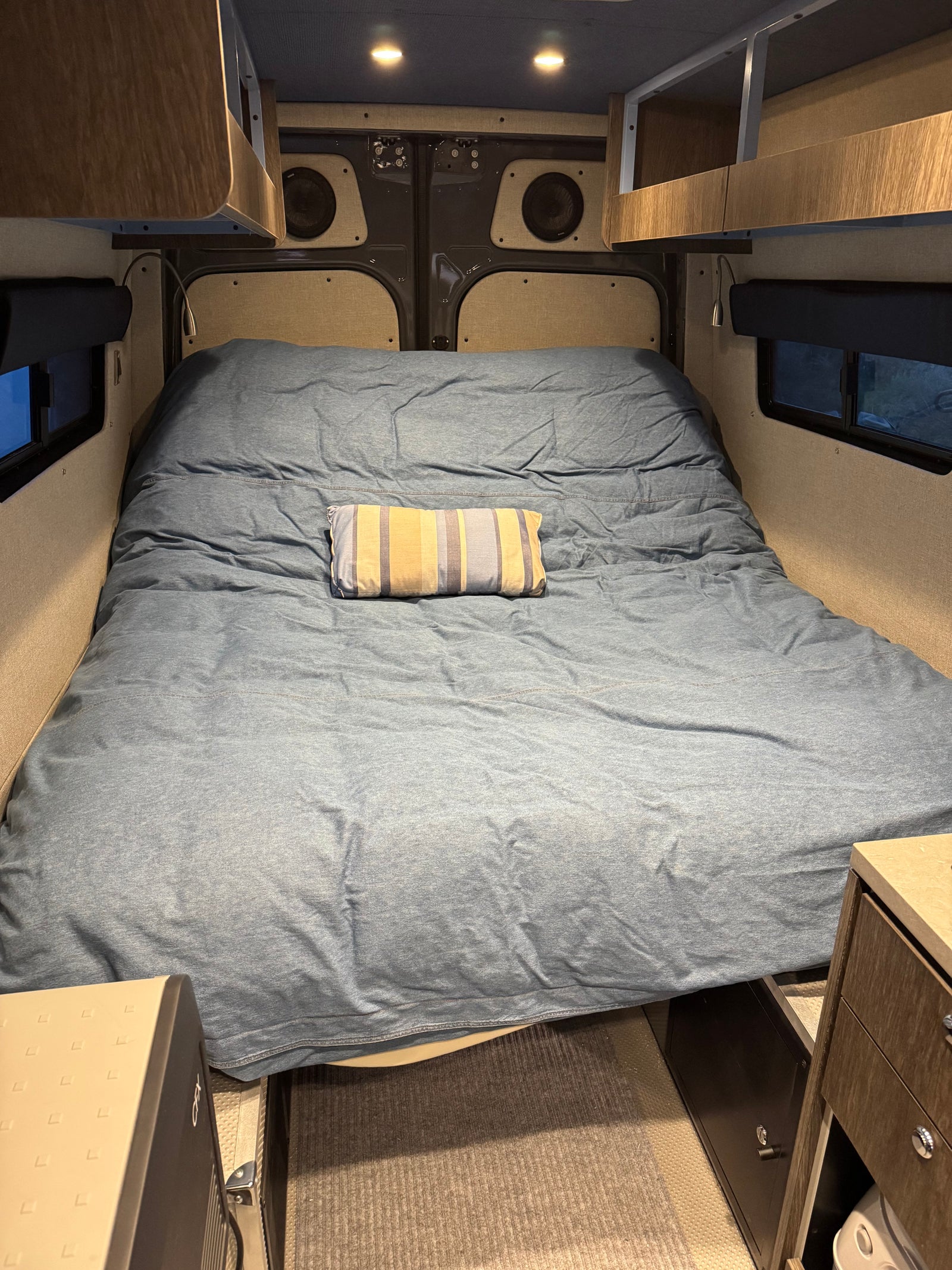 Cozy bed area in an RV rental with blue bedding, overhead storage, speakers, and under-bed appliances and safe.