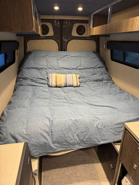 Cozy bed area in an RV rental with blue bedding, overhead storage, speakers, and under-bed appliances and safe.