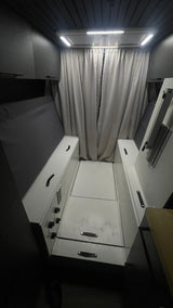 Compact RV rental interior: A narrow aisle between a fold-up bed & storage, illuminated by LED lights and curtains.