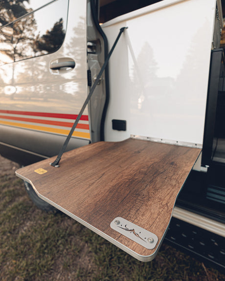 Outdoor culinary setup on an RV rental: a wood-grain fold-out table supported by a strap, with custom mountain art.