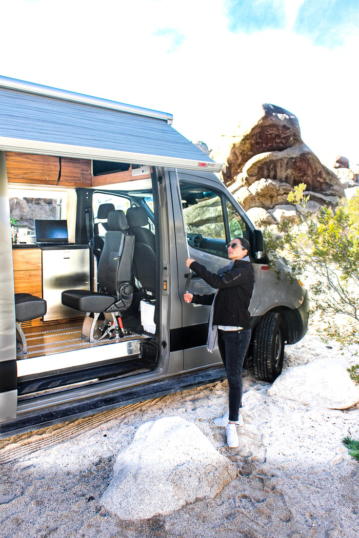 A woman prepares her RV rental, extending the awning outside a grey camper van in a rocky desert. Interior shows kitchen and seating.