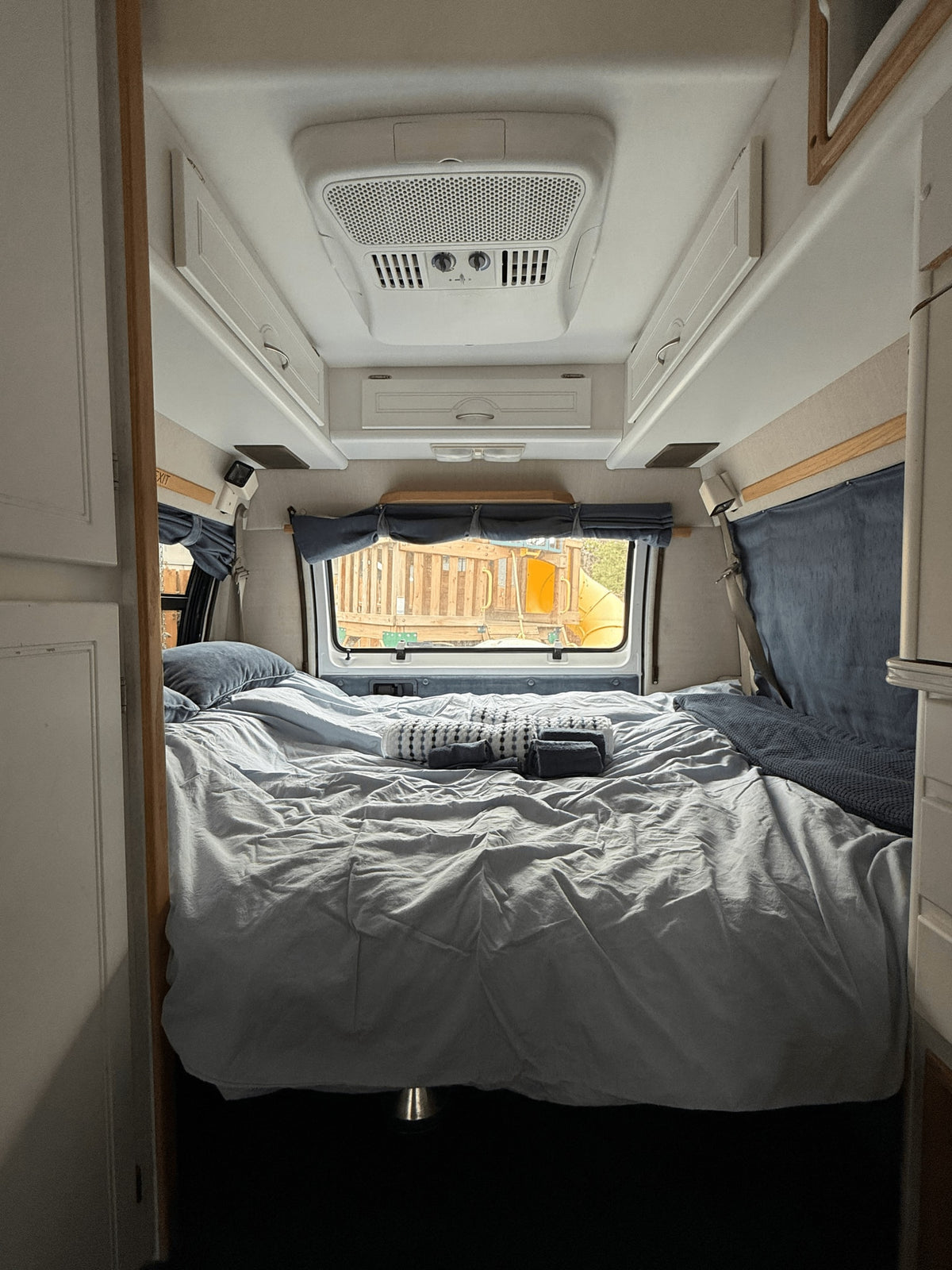An RV rental's cozy sleeping area with a made bed, overhead AC, storage, and a playground outside the rear window.
