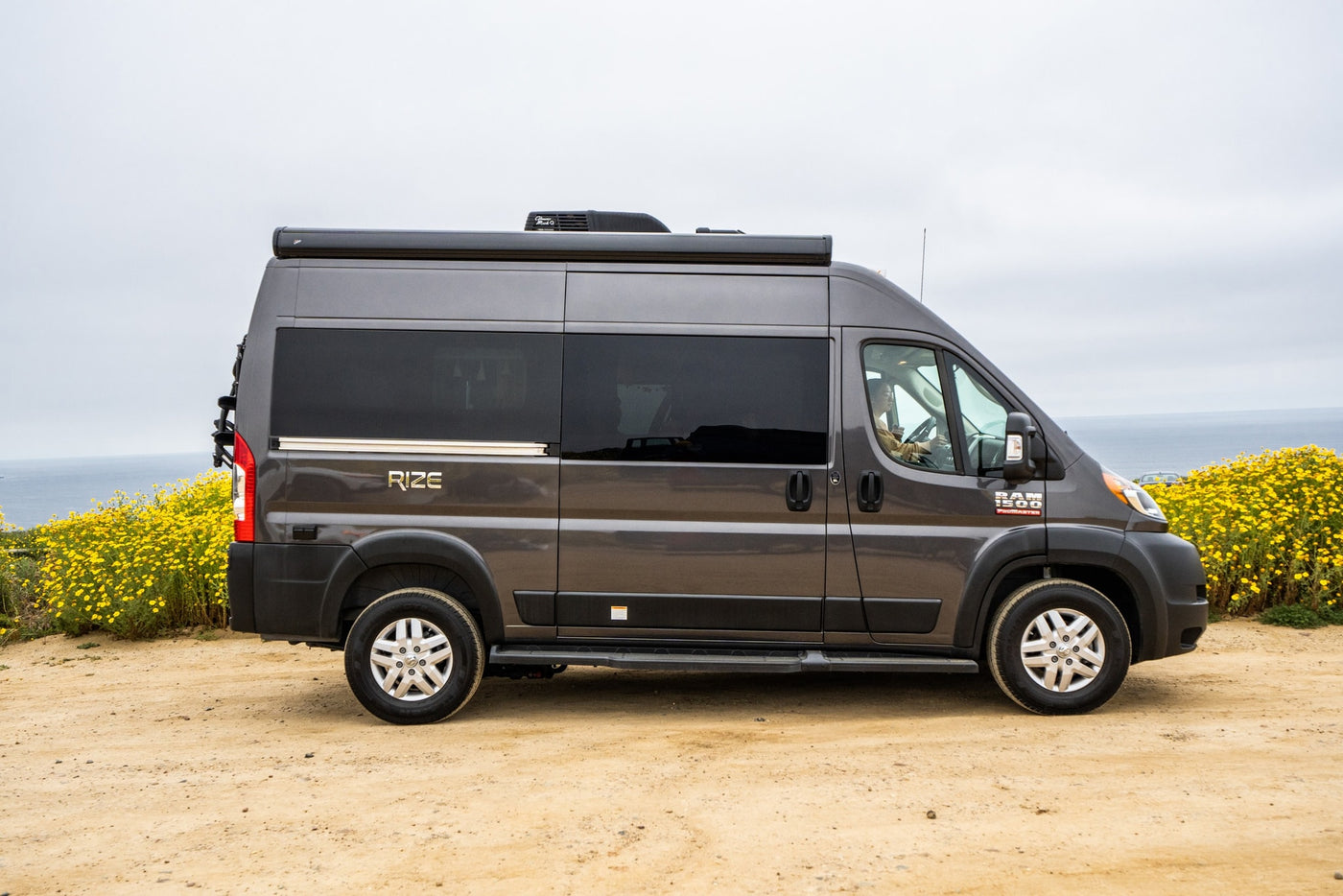 This gray RIZE Ram ProMaster RV rental is parked on a dirt road with yellow flowers and the ocean in background. — photo 1