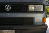 Front of a blue-grey VW van, ideal for RV rental. Close-up: central VW emblem on black grille, dual headlights.