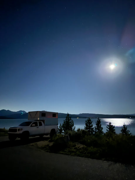 RV rental truck camper parked lakeside under a bright moon and stars. Serene night camping with mountains in view.